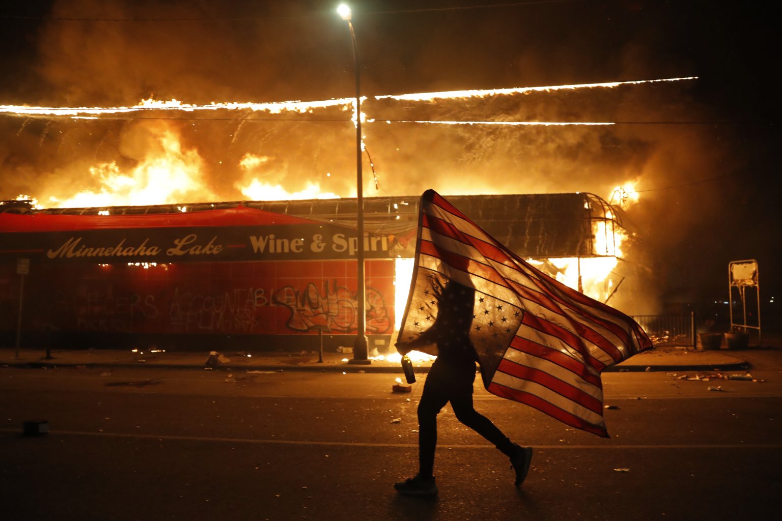 Demonštrant nesie americkú vlajku vedľa horiacej budovy počas pokračujúcich protestov proti policajnému zásahu v americkom meste Minneapolis vo štvrtok 28. mája 2020. Foto: TASR/AP
