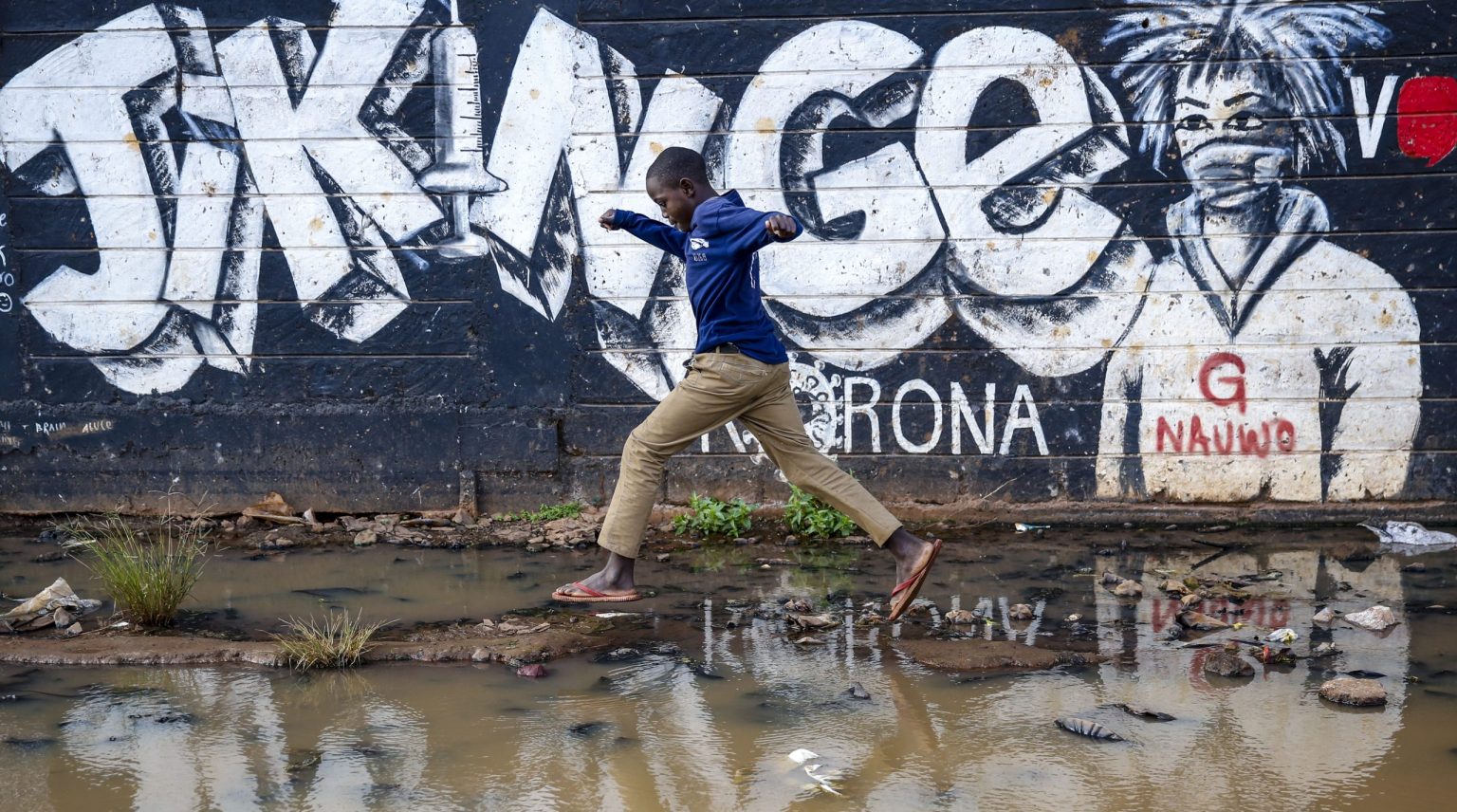 Chlapec kráča po zaplavenej ulici okolo nápisu, ktorý vyzýva ľudí, aby sa dali očkovať proti ochoreniu COVID-19 v chudobnej štvrti Kibera v Nairobi. Foto: TASR/AP